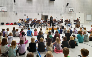 Orkesteri soittamassa koulun salissa, jossa istuu lapsia.